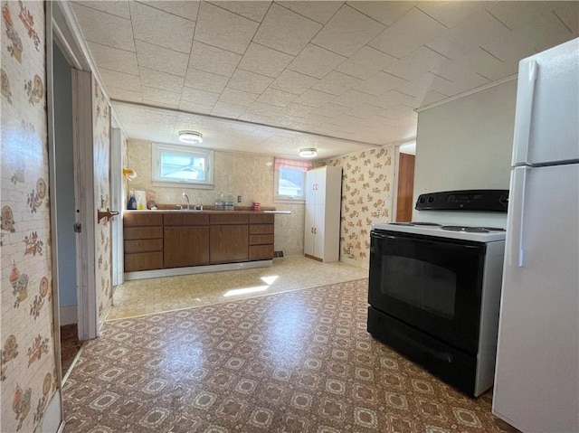 kitchen with black range with electric stovetop, a sink, freestanding refrigerator, brown cabinetry, and wallpapered walls