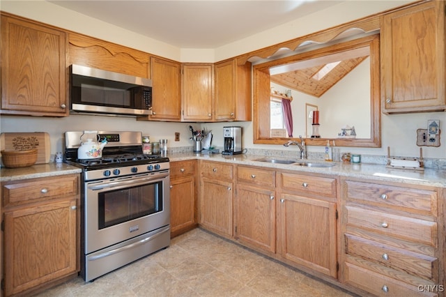 kitchen featuring light stone counters, brown cabinetry, vaulted ceiling with skylight, a sink, and appliances with stainless steel finishes