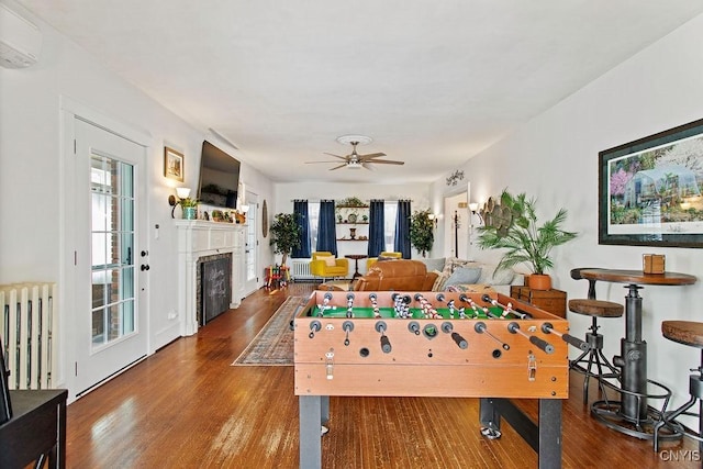 playroom featuring wood finished floors, a wall mounted AC, radiator heating unit, a fireplace, and ceiling fan