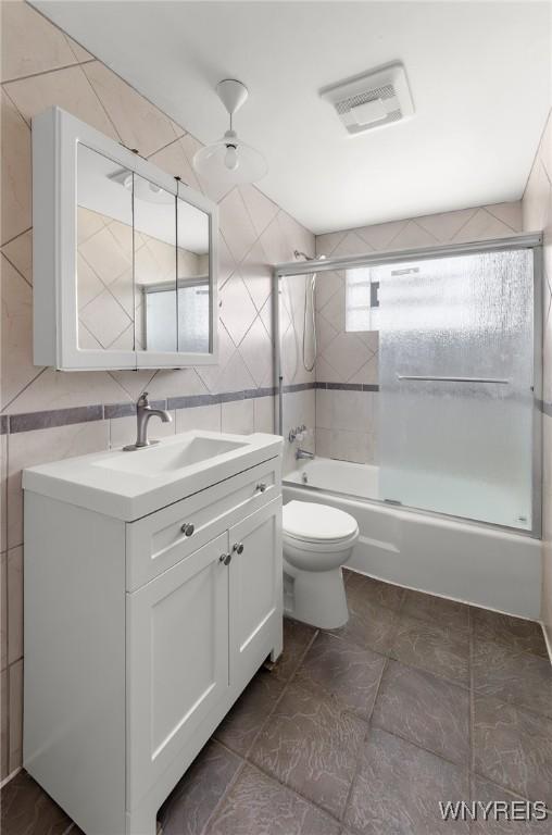 bathroom featuring visible vents, toilet, tile walls, bath / shower combo with glass door, and vanity