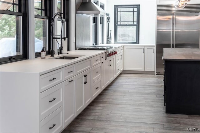 kitchen featuring light wood finished floors, wall chimney range hood, light countertops, appliances with stainless steel finishes, and a sink