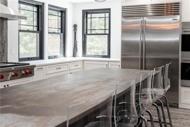 kitchen with white cabinetry, appliances with stainless steel finishes, wall chimney exhaust hood, light wood finished floors, and light stone countertops