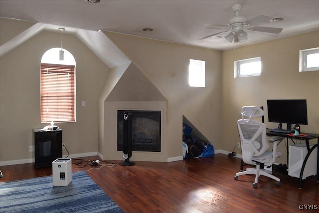 office space featuring baseboards, vaulted ceiling, wood finished floors, a glass covered fireplace, and a ceiling fan