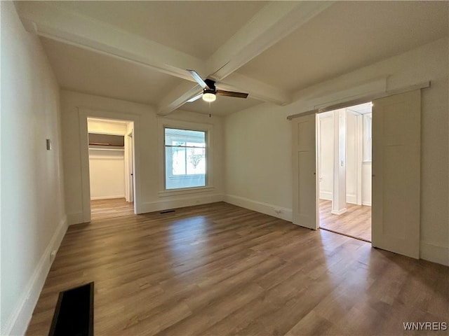 unfurnished bedroom featuring wood finished floors, baseboards, ensuite bath, a spacious closet, and beamed ceiling