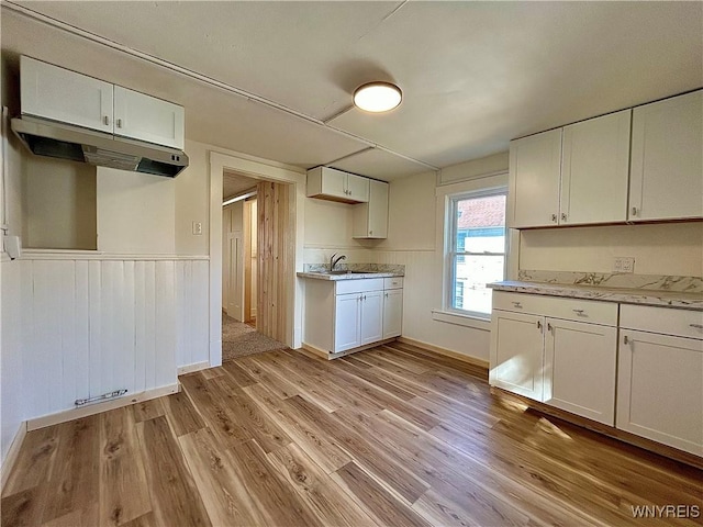 kitchen with light wood-style flooring, a sink, white cabinets, under cabinet range hood, and wainscoting