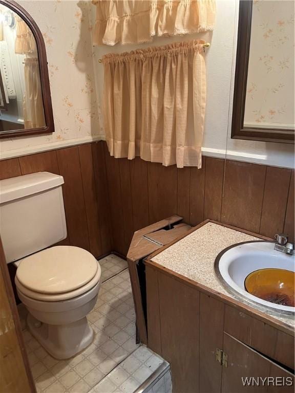 half bathroom with vanity, a wainscoted wall, wallpapered walls, tile patterned floors, and toilet