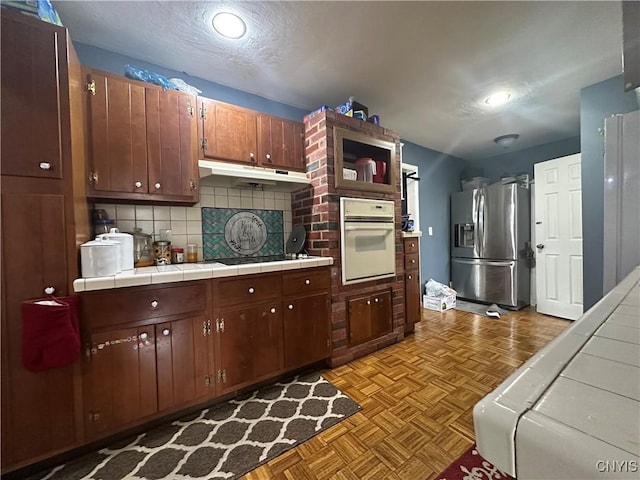 kitchen featuring tasteful backsplash, oven, tile counters, under cabinet range hood, and stainless steel fridge