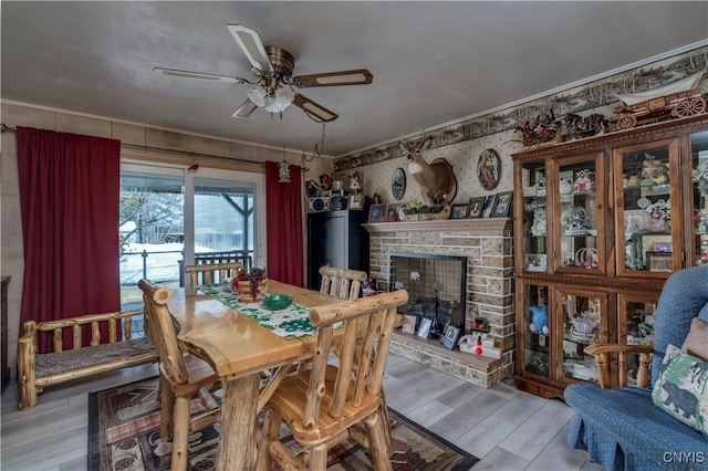 dining room with light wood-type flooring, ornamental molding, a ceiling fan, a stone fireplace, and wallpapered walls