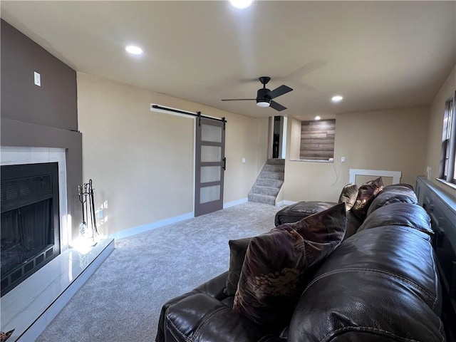 living area featuring stairway, a ceiling fan, a fireplace with raised hearth, a barn door, and carpet flooring