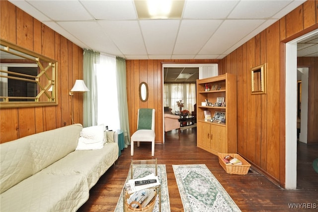 living room featuring a drop ceiling, wooden walls, and dark wood finished floors