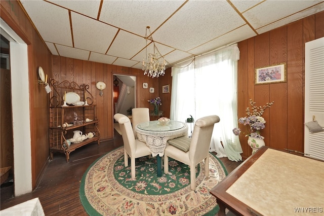 dining space with dark wood finished floors, an inviting chandelier, a drop ceiling, and wood walls