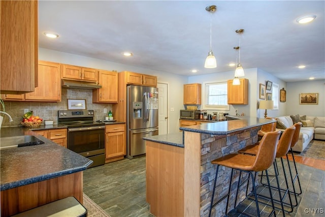 kitchen with a sink, under cabinet range hood, a kitchen breakfast bar, appliances with stainless steel finishes, and decorative backsplash