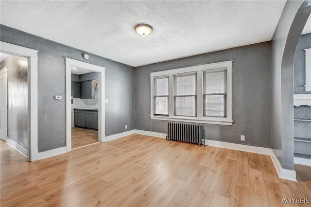 unfurnished bedroom featuring radiator, baseboards, light wood-style flooring, arched walkways, and a textured ceiling