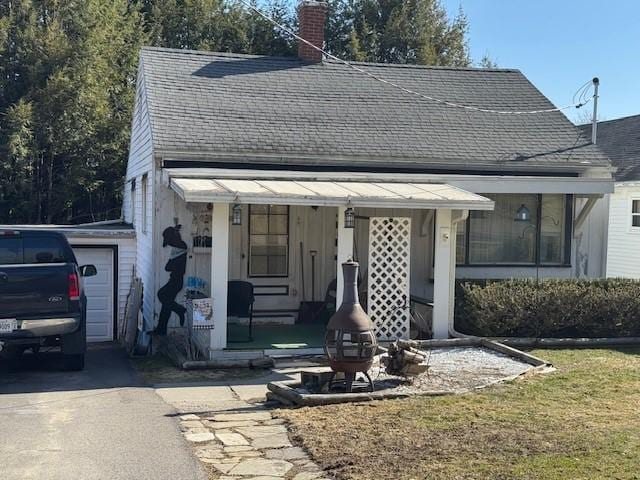 view of front facade with a shingled roof, covered porch, a chimney, a garage, and driveway