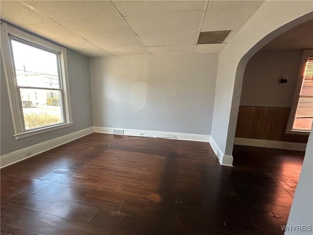 unfurnished room with wood finished floors, arched walkways, a drop ceiling, and baseboards