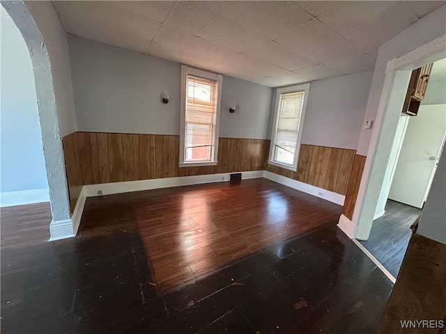 unfurnished room with a drop ceiling, wood finished floors, arched walkways, wood walls, and wainscoting