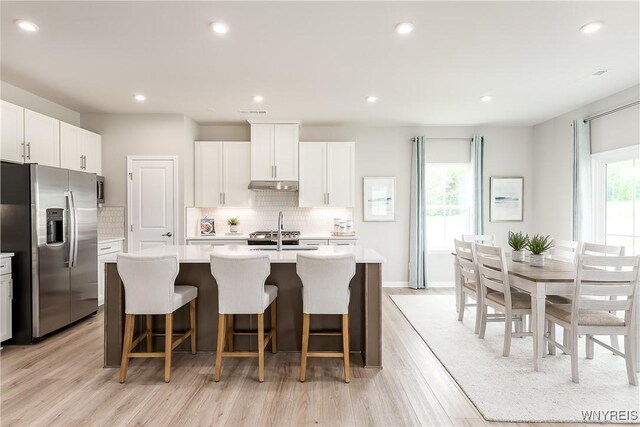 kitchen with backsplash, light countertops, an island with sink, a wealth of natural light, and stainless steel refrigerator with ice dispenser