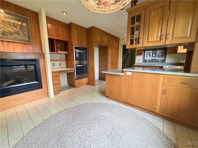 kitchen featuring double oven, built in desk, brown cabinetry, a glass covered fireplace, and a sink