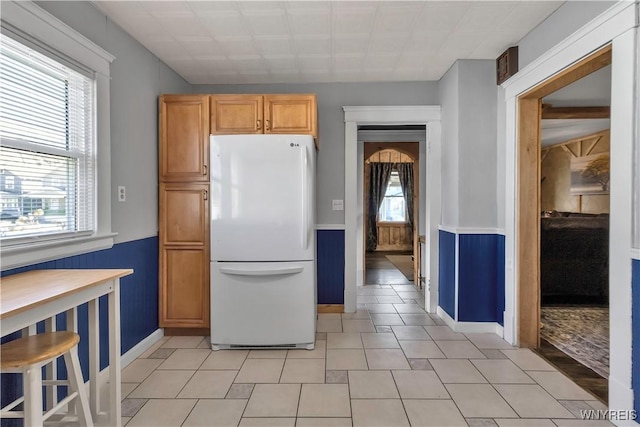 kitchen featuring freestanding refrigerator, baseboards, light tile patterned floors, and a wainscoted wall