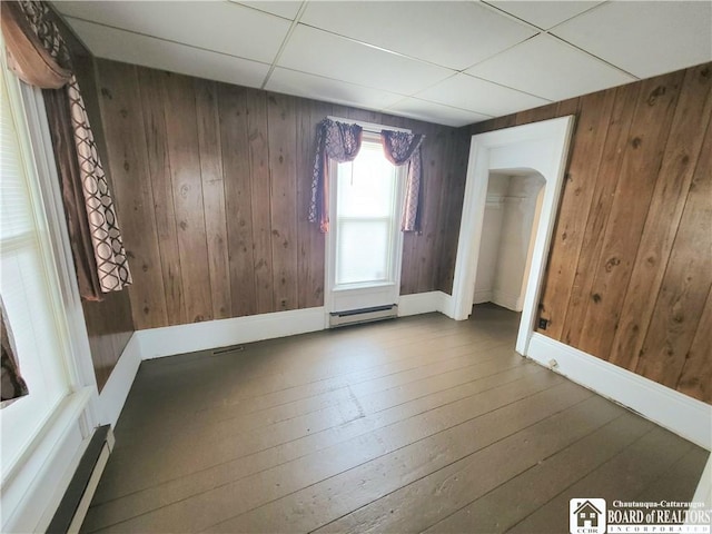 unfurnished room featuring wooden walls, hardwood / wood-style floors, baseboards, a baseboard heating unit, and a drop ceiling