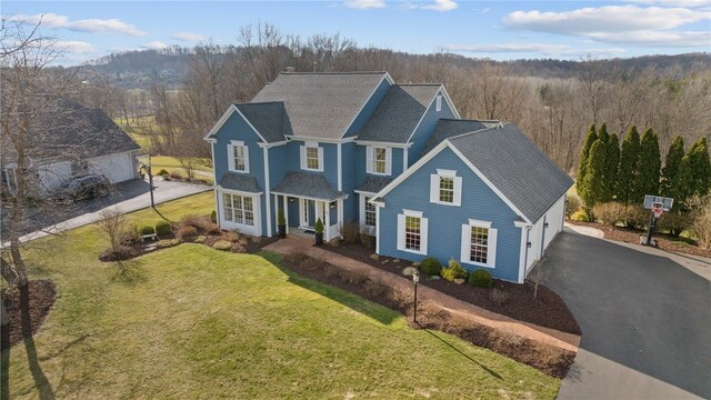 traditional home featuring driveway, a wooded view, roof with shingles, covered porch, and a front lawn