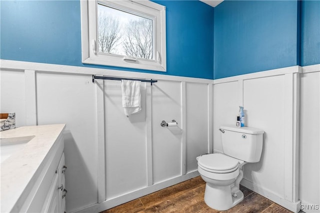 half bathroom with a wainscoted wall, toilet, vanity, wood finished floors, and a decorative wall
