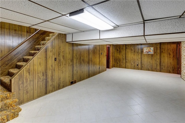 finished basement featuring visible vents, wooden walls, stairs, tile patterned floors, and a drop ceiling