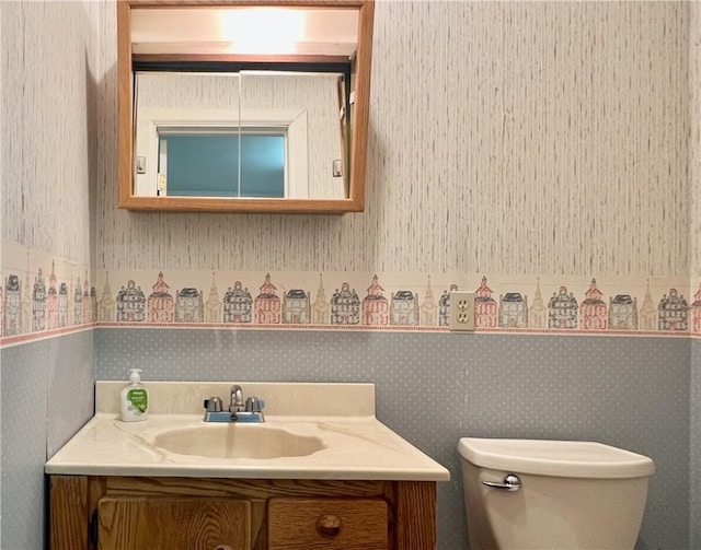 half bath featuring a wainscoted wall, wallpapered walls, toilet, and vanity