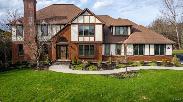 tudor house featuring a chimney, a front yard, brick siding, and roof with shingles