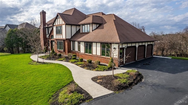 english style home featuring aphalt driveway, a chimney, a garage, a front yard, and brick siding