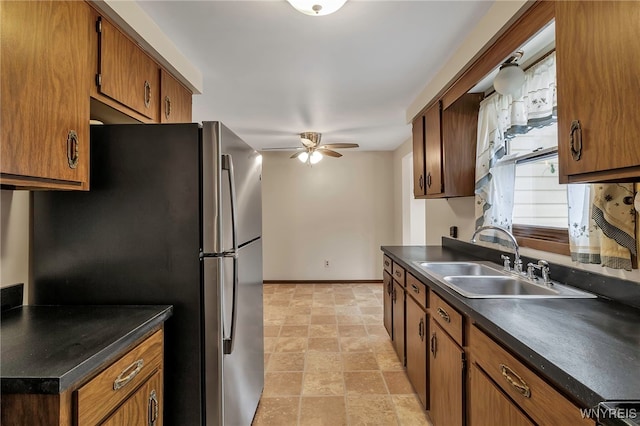 kitchen featuring ceiling fan, freestanding refrigerator, brown cabinetry, a sink, and dark countertops