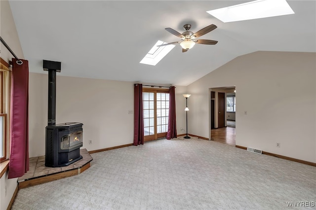 unfurnished living room with vaulted ceiling with skylight, a wood stove, a ceiling fan, visible vents, and carpet flooring