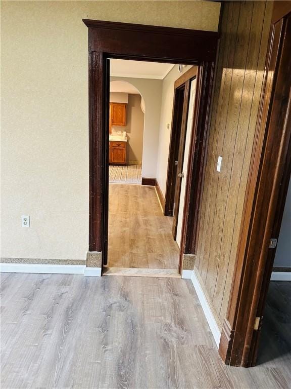 corridor featuring baseboards, ornamental molding, wood walls, arched walkways, and light wood finished floors