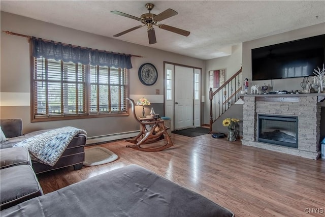 living area with ceiling fan, stairs, a stone fireplace, baseboard heating, and wood finished floors