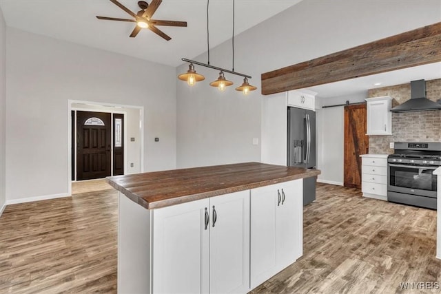 kitchen with butcher block counters, wall chimney exhaust hood, ceiling fan, stainless steel appliances, and wood finished floors