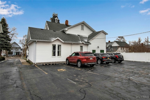 exterior space with uncovered parking, roof with shingles, a chimney, and fence