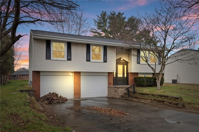 raised ranch featuring roof with shingles, driveway, an attached garage, brick siding, and a lawn