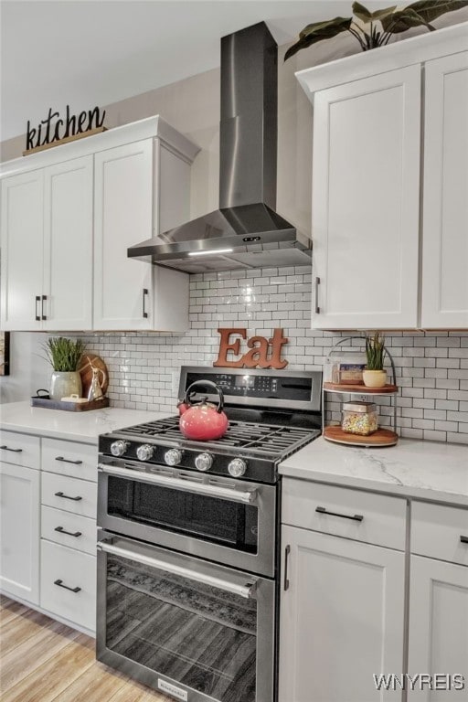 kitchen with white cabinets, light wood-type flooring, wall chimney exhaust hood, double oven range, and tasteful backsplash