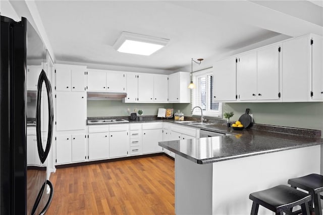 kitchen featuring under cabinet range hood, dark countertops, a sink, freestanding refrigerator, and a peninsula