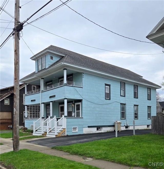 traditional style home with a balcony, a front lawn, a shingled roof, covered porch, and fence