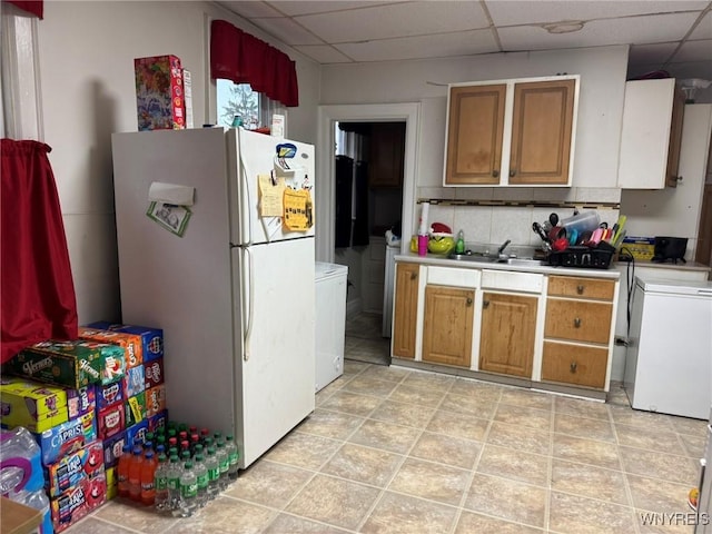 kitchen with a sink, brown cabinets, light countertops, a paneled ceiling, and freestanding refrigerator