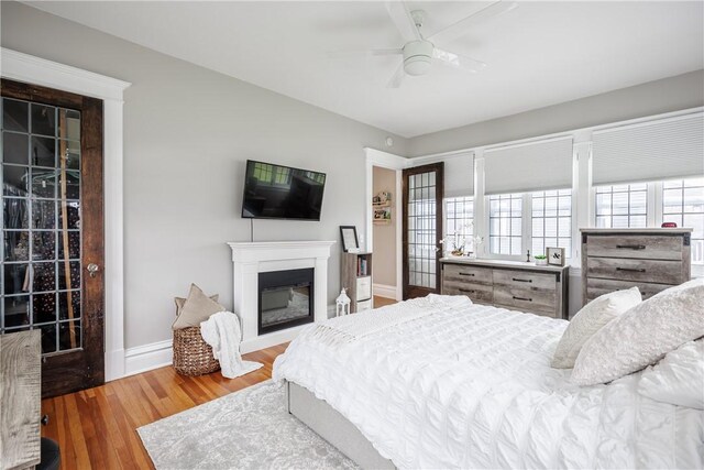 bedroom featuring a glass covered fireplace, wood finished floors, baseboards, and ceiling fan