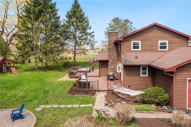 back of property featuring a playground, a chimney, a lawn, a wooden deck, and roof with shingles