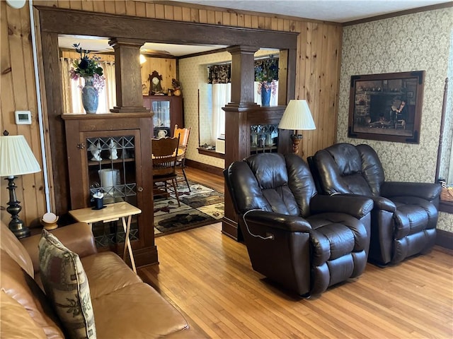 living room featuring wallpapered walls, wood finished floors, crown molding, and decorative columns