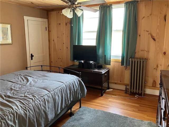 bedroom featuring light wood-style flooring, baseboards, radiator heating unit, ceiling fan, and wooden walls