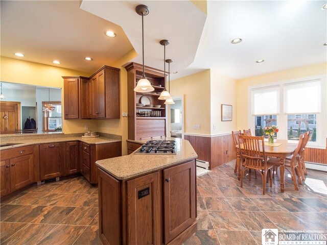 kitchen featuring baseboard heating, stainless steel gas cooktop, brown cabinetry, wainscoting, and light stone countertops