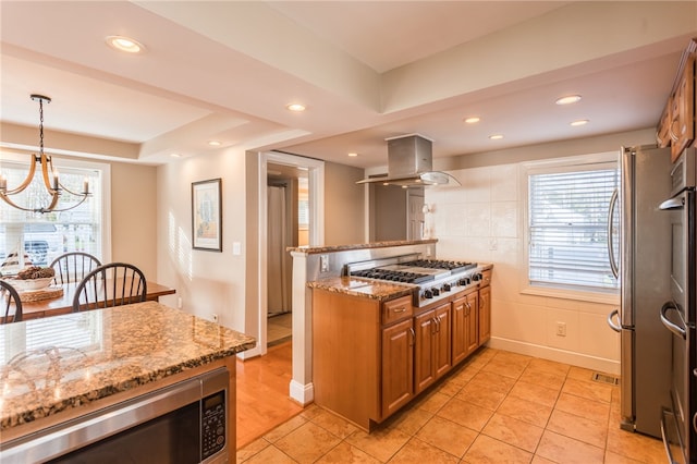kitchen with island exhaust hood, light stone counters, brown cabinetry, appliances with stainless steel finishes, and a notable chandelier