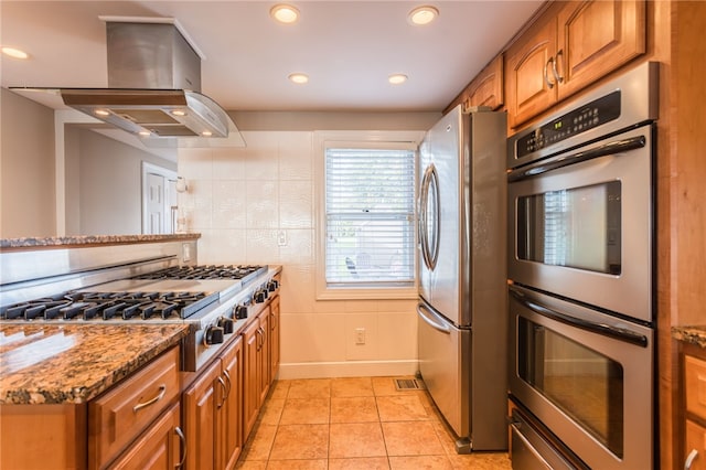 kitchen featuring light stone counters, brown cabinetry, light tile patterned flooring, island exhaust hood, and appliances with stainless steel finishes