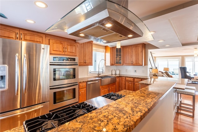 kitchen with island exhaust hood, a sink, brown cabinets, light stone counters, and appliances with stainless steel finishes