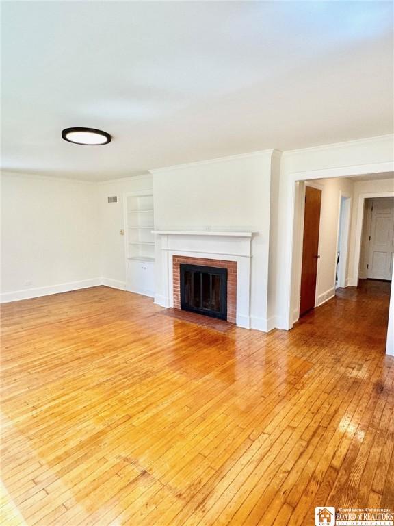 unfurnished living room with crown molding, baseboards, light wood-style flooring, a brick fireplace, and built in features